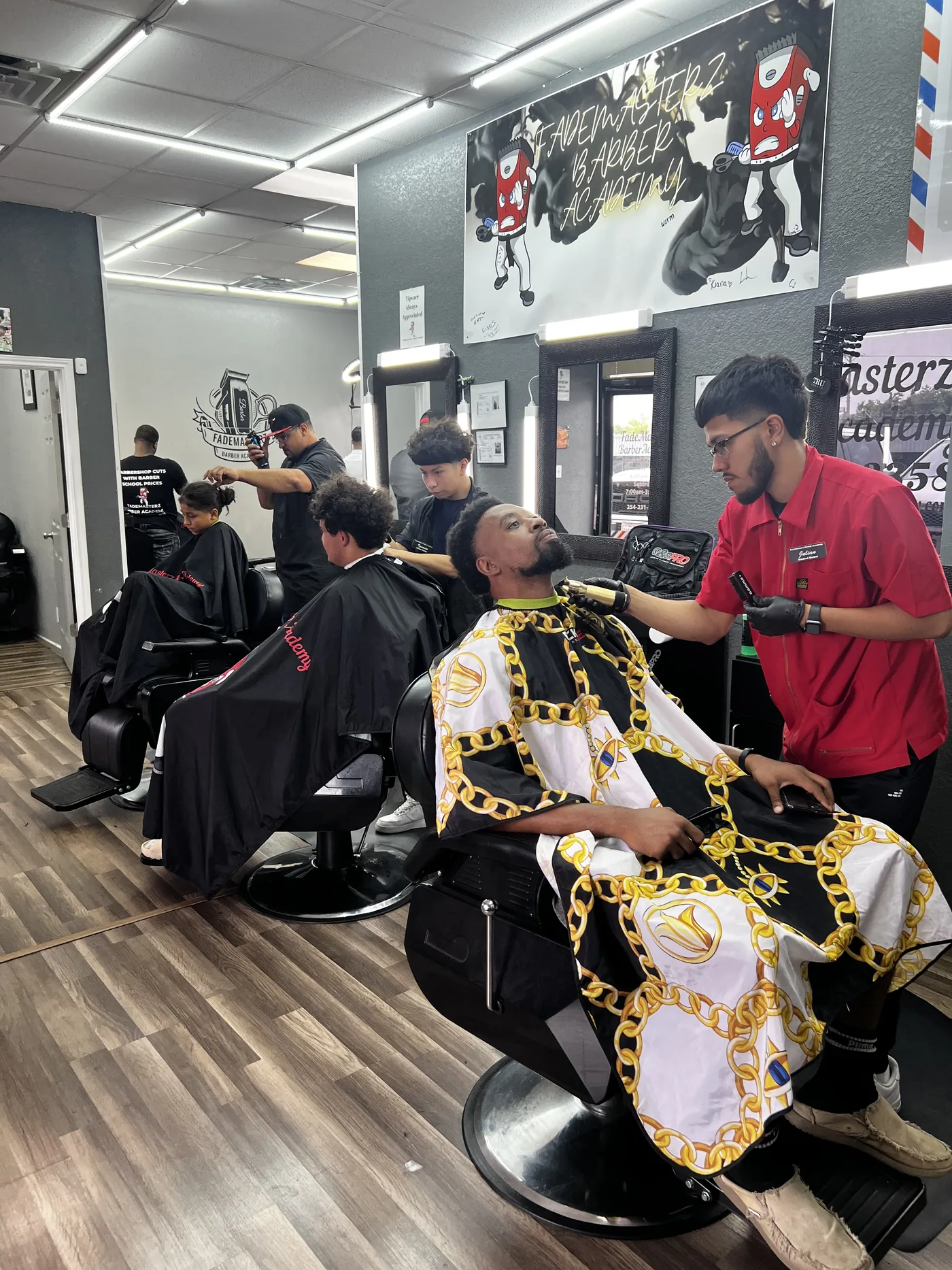 A student cuts a client in a gold-chain cape under the FadeMasterz mural